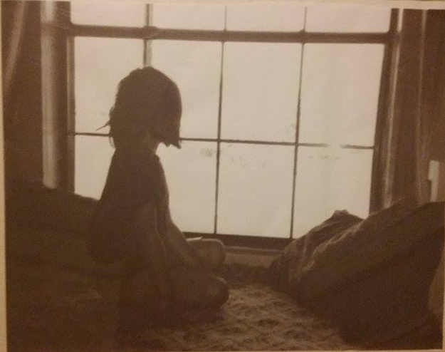 Photograph in sepia tones of a young girl looking out a bay window she is seated at.